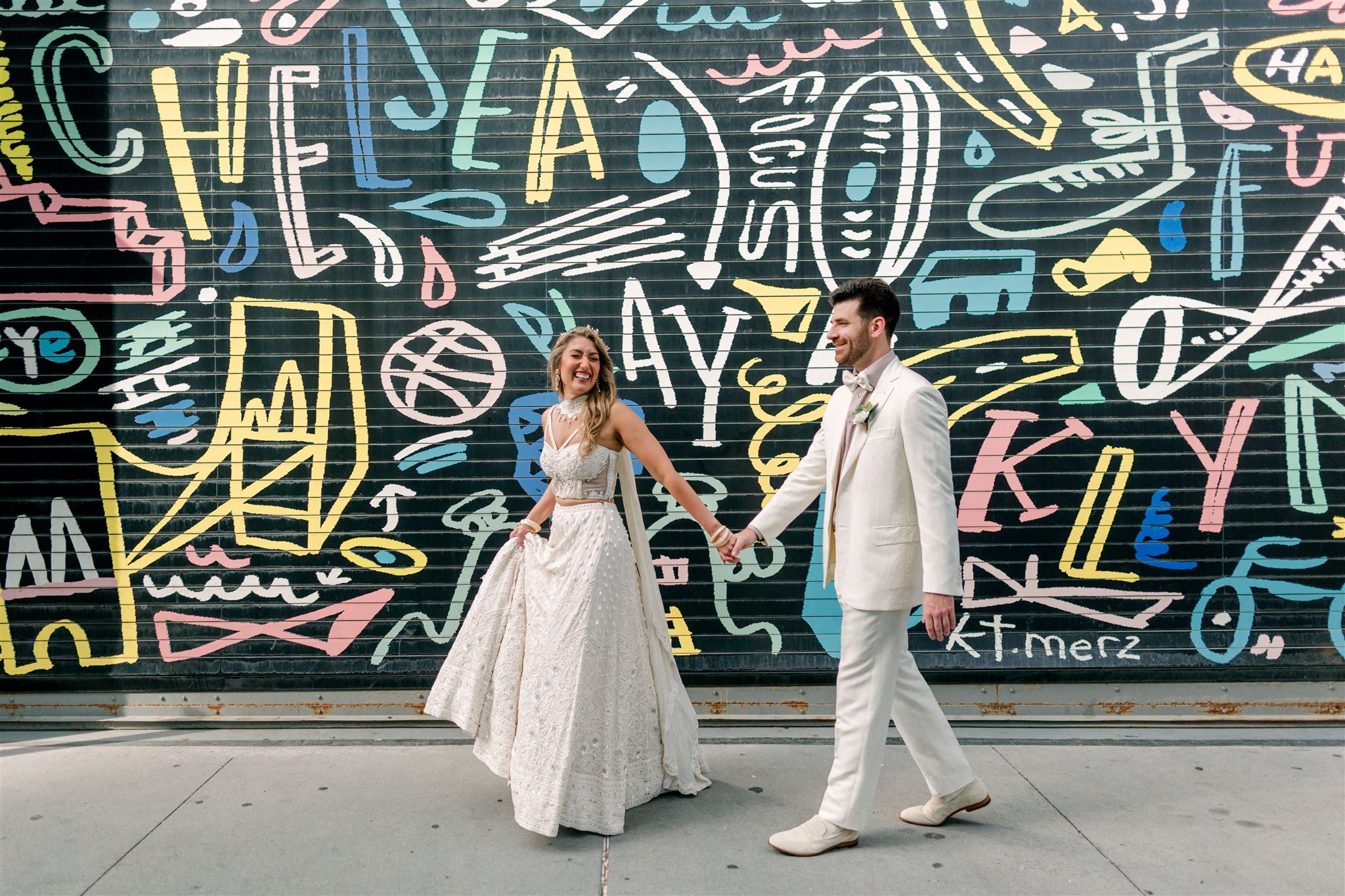 Bride and groom in white walking hand in hand in front of a colorful mural in New York City on their wedding day