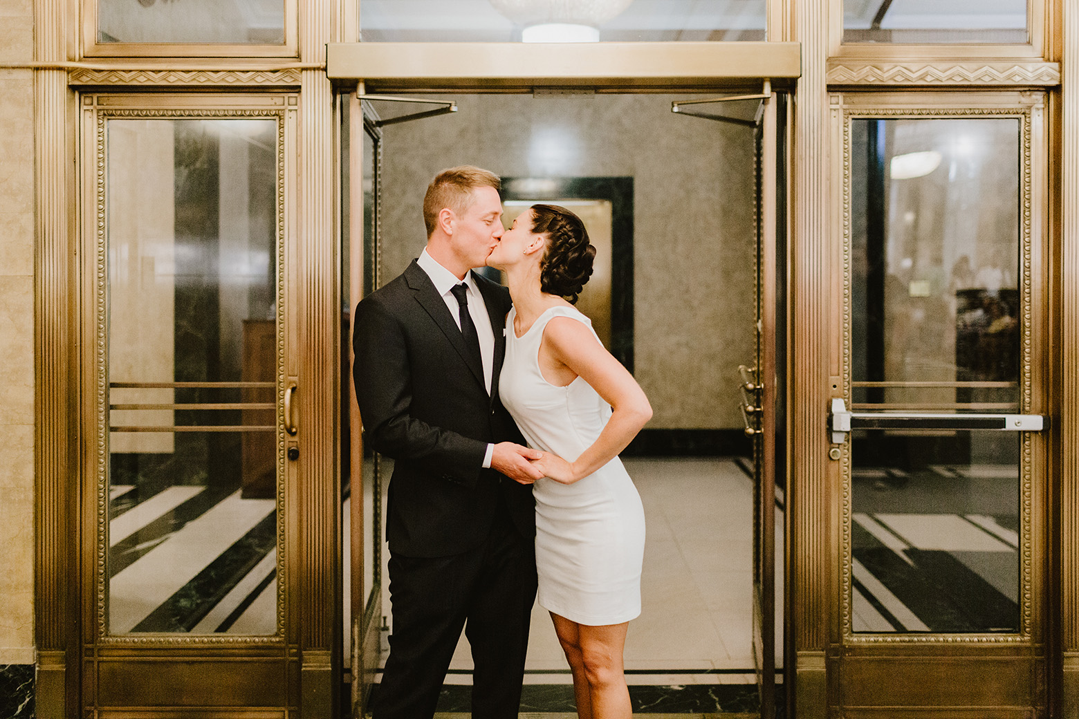 a woman kissing a man in NYC City Hall. The woman is wearing a short white dress while the man is wearing a black suit for their NYC elopement
