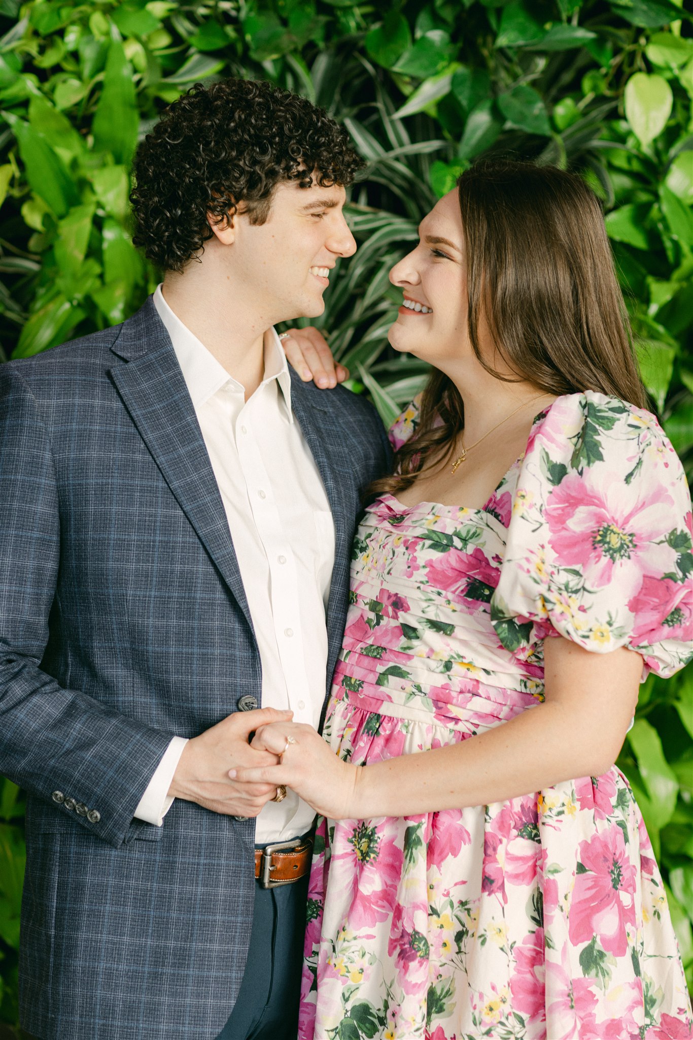 A couple holding hands, the girl is wearing a pink floral dress and the man is wearing a suit during their engagement session