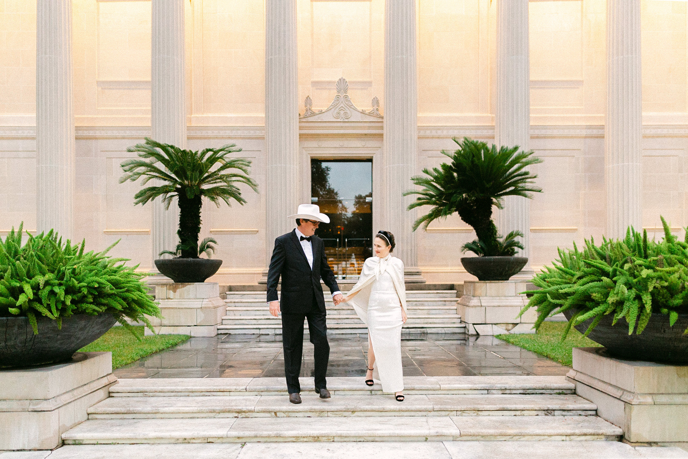 A bride and groom holding hands walking outside of their Houston wedding venue. The groom is wearing a white cowboy hat with his tux