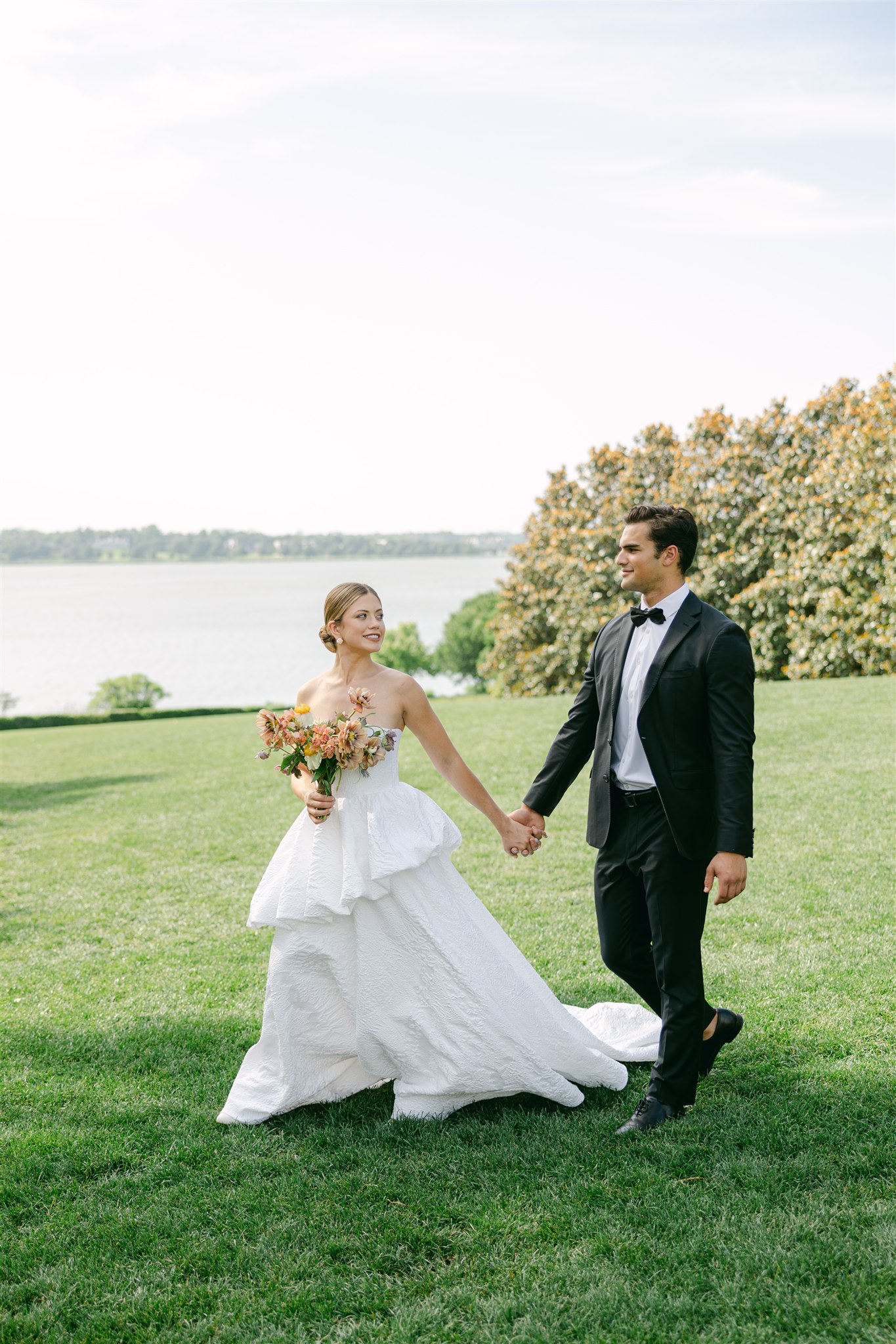 a wedding couple walking hand in hand across the lawn at the dallas arboretum with white rock lake in the background