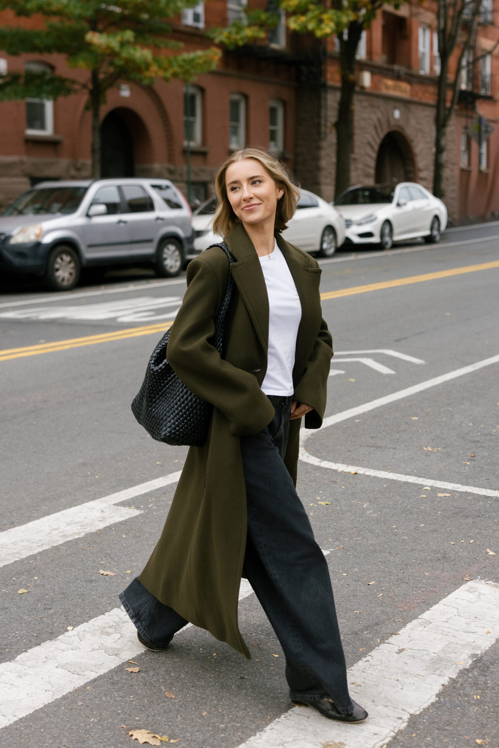 a girl wearing an olive green trench coat walking across the street in nyc