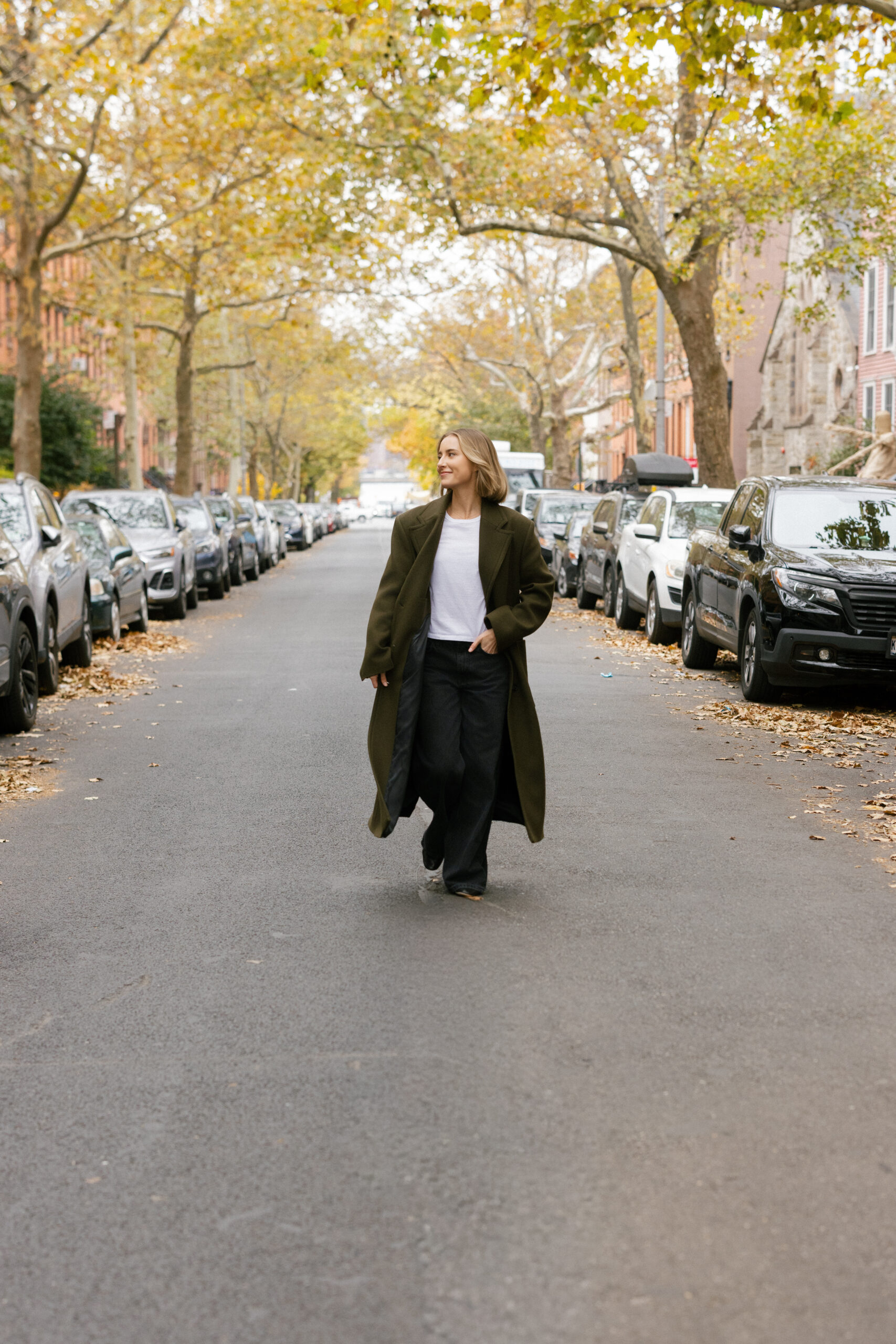 a girl wearing an olive green trench coat walking across the street in nyc