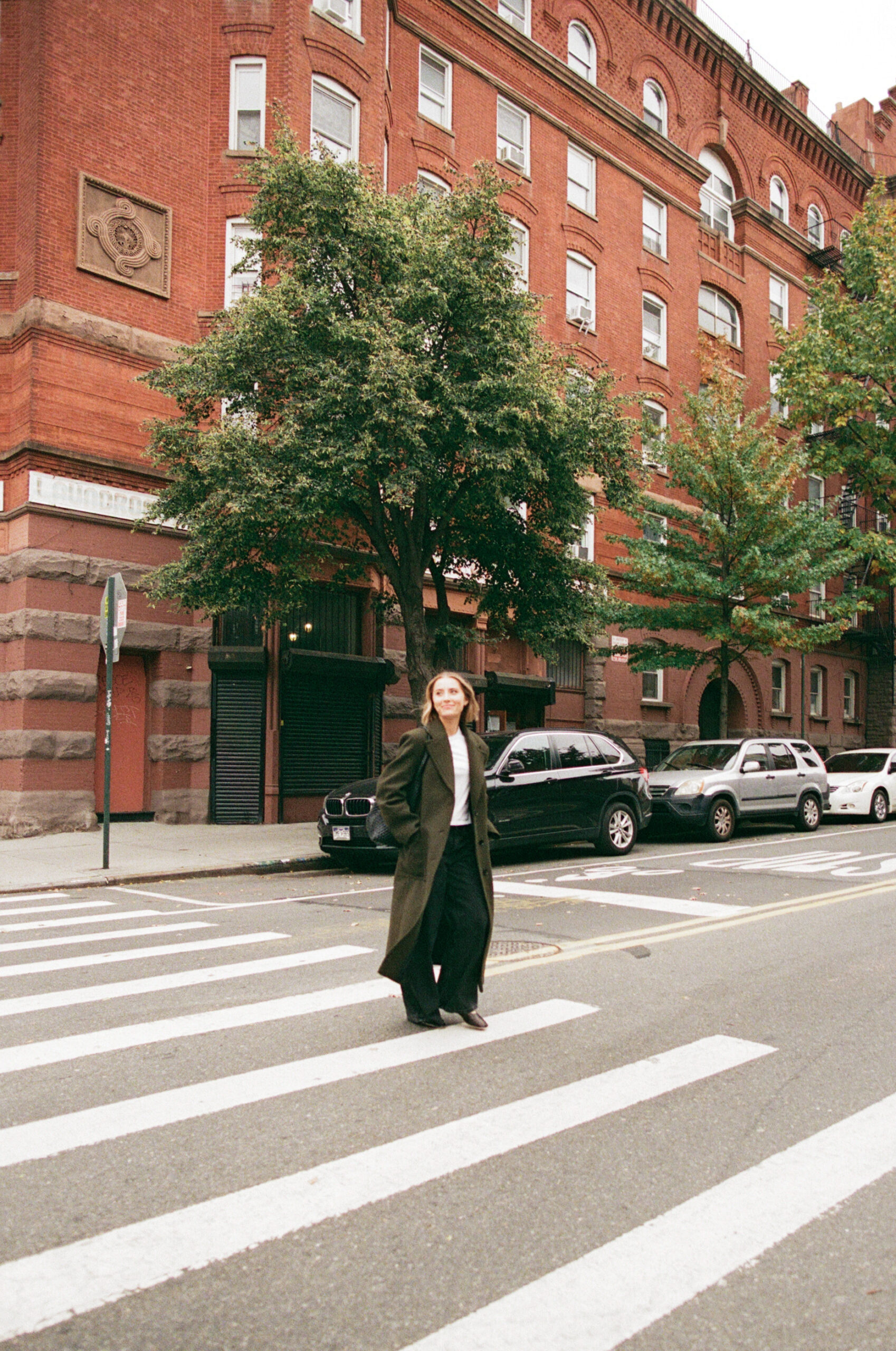 a girl wearing an olive green trench coat walking across the street in nyc