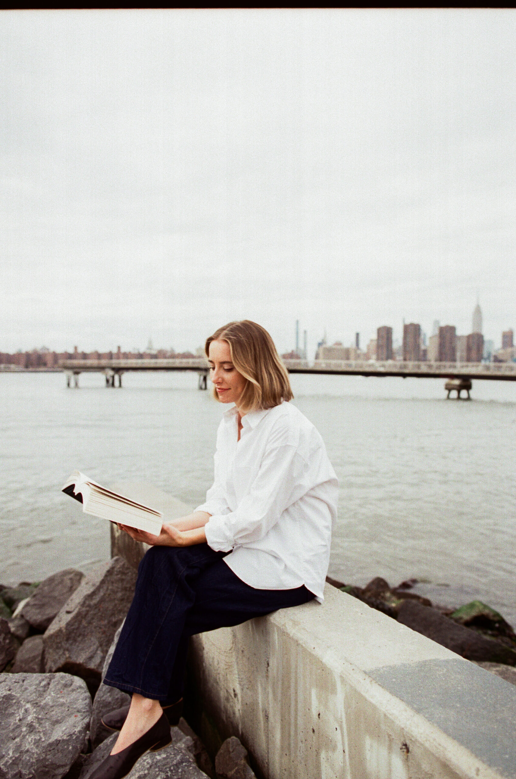 a girl wearing a white button down in front of the ocean with nyc skyline in the background
